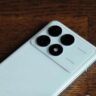 Redmi K70 Pro Price In India: A white Redmi K70 Pro smartphone with a circular camera module featuring four lenses and a flash, resting on a wooden surface.